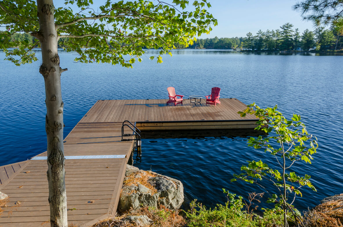 Looking out onto a lake with an L-shaped dock and two red adirondack chairs. 